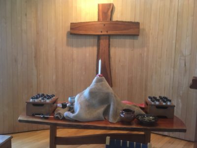 single candle in child's clay holder elevated above altar table, draped in burlap